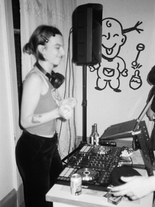 black and white photo of izzy djing. there is a desk in front of her with DJ decks, bottles of drinks and a laptop on it. There is a speaker in the background and paintings on the wall