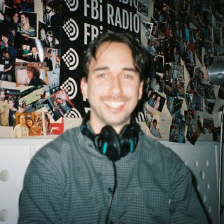 Radio presenter Ben Hansen sits in front of a wall of photos and an fbi radio sign. Film photo. Blurry.