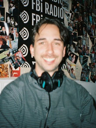 Radio presenter Ben Hansen sits in front of a wall of photos and an fbi radio sign. Film photo. Blurry.