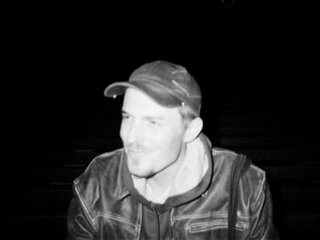 A black and white photo of John Troughton wearing a seated, wearing a jacket and looking to the left of the frame.