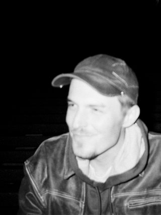 A black and white photo of John Troughton wearing a seated, wearing a jacket and looking to the left of the frame.