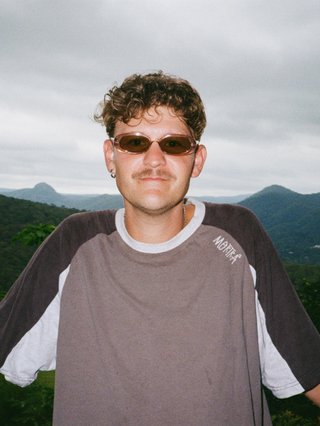 Harrie Hastings stands in front of cloud-covered sky and rolling hills. He's wearing sunglasses and a t-shirt.