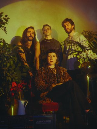 An image of four members of the band Kyoshi standing amidst green foliage. A band member is seated while three others stand behind them, with a yellow light illuminating the background.