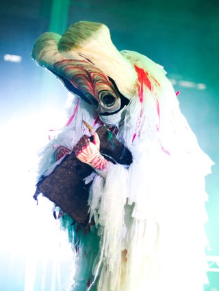 Hunched figure draped in woven whites, beiges, and a hint of orange. Against a green hue backgrounf