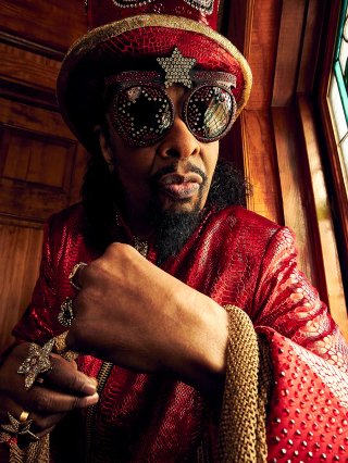 Bootsy Collins is staring into camera in a wooden room with a stained glass window to his right. he is holding a fist up to the camera and is wearing bedazzled glasses