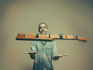 Larry Crestani of Standing Circle standing before a light green, shadowed wall wearing a blue button up shirt holding his arms out with palms faced upward; his kraartar instrument floating before his face.