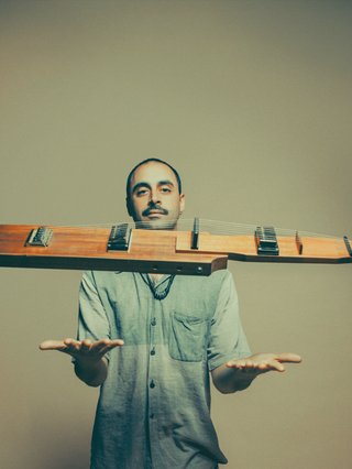 Larry Crestani of Standing Circle standing before a light green, shadowed wall wearing a blue button up shirt holding his arms out with palms faced upward; his kraartar instrument floating before his face.