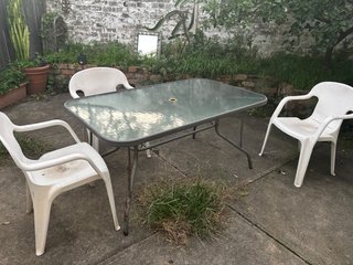 Three white plastic chairs surround a glossy grey-ish blue table in a concrete backyard. Weeds and plants grow from in between the cracks and in a garden at the back of the picture.
