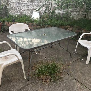 Three white plastic chairs surround a glossy grey-ish blue table in a concrete backyard. Weeds and plants grow from in between the cracks and in a garden at the back of the picture.