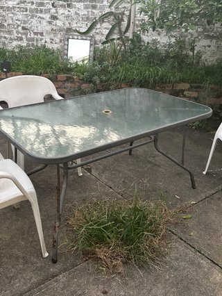 Three white plastic chairs surround a glossy grey-ish blue table in a concrete backyard. Weeds and plants grow from in between the cracks and in a garden at the back of the picture.
