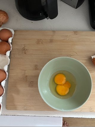 photo of a wooden cutting board. a green bowl is resting on it with two egg yolks in it. on the left there is a 12 pack of eggs with one cracked egg shell on the side. you see the corner of a kettle at the top of the image too