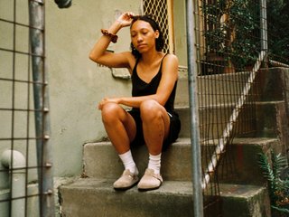 Bri is sitting on a stoop of a house touching her head. She is wearing a black singlet and shorts.