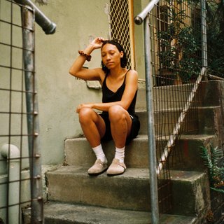 Bri is sitting on a stoop of a house touching her head. She is wearing a black singlet and shorts.