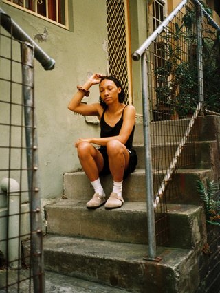 Bri is sitting on a stoop of a house touching her head. She is wearing a black singlet and shorts.
