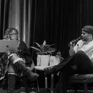 Ify Obeigbu (left) sits on a chair facing the audience with her face turned towards Tasman Keith (right) who holds a microphone speaking towards the audience but his head is turned to Ify. They sit in front of a black curtain background and the image is in grainy black and white.