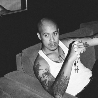 Black and white image of BAYANG sitting on a couch. He is wearing a white singlet and black pants. He is holding rosary beads.