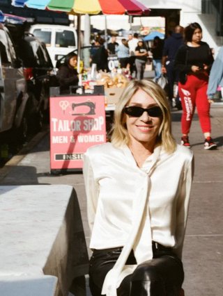 Kim Gordon wearing sunglasses sits on a chair on a street sidewalk. A tailor shop sign and people can be seen in the background