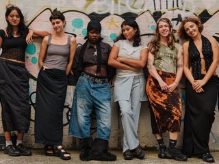 The six hosts of Dance Class stand against a brick wall with graffiti on it. They are all excited.
