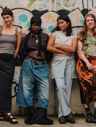 The six hosts of Dance Class stand against a brick wall with graffiti on it. They are all excited.