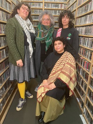 Three people, two wearing keffiyehs, stand in fbi.radio's music library. In front of them, another person is seated with a shawl draped over them.