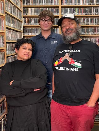 Shareeka, Ethan and Rosco stand in the fbi music library. They look into the camera directly. Rosco's short shws two hands comign together with the text BLACKFULLA PALESTINIANS in solidarity