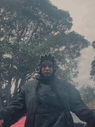 Shareeka stands in front of the camera on a gloomy day, there's a red car and a tree in the background. She's wearing a big black puffer and a knitted bonnet