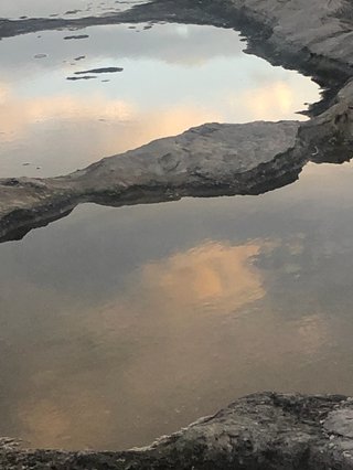A sandstone rock on the coast with puddles of ocean water. The water is clear, and it's reflecting the sky as though the clouds at sunset