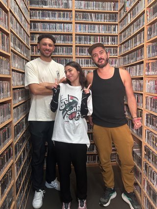 Three people stand in the fbiradio CD archive library. From left to right is Ahmad Khochaiche, Sara El Youghun, and Maxime Camo. Ahmad wears a white t-shirt, dark jeans and white sneakers. Sara wears a white t-shirt with a Palestinian flag displayed, and black pants. Maxime wears a black tank top and camel coloured pants.