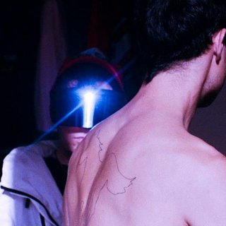 Photo of both members of the band Collarbones. One of the members is wearing a shiny visor that covers his eyes and us facing the camera. The other member is shirtless and facing away with his back to the camera.