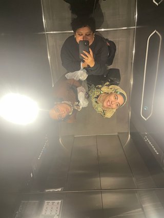 Toobs, Shareeka and Sara stand in a lift together. There's a mirror on a ceiling, where they're looking up to capturing this selfie