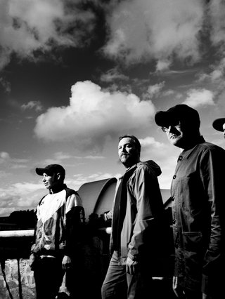 4-piece Scottish band Mogwai pose standing in the right of frame. Fluffy clouds make up the top 2/3 of the image. The image is in greyscale.