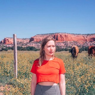 Julia Jacklin stands in a field of yellow flowers, with rocky orange mountains behind her. She is looking slightly to the right of the camera with a slightly concerned expression.