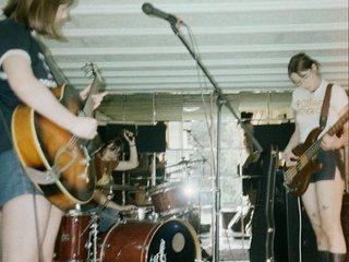 Sevilles band playing a show, guitarist to the front left (facing the image) drummer in the back, bass player right back (facing the image)