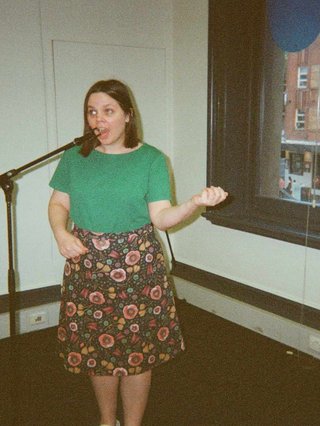Bre wearing a green top tucked into a floral knee-length skirt and standing in front of an empty mic stand being very silly and miming playing guitar and singing