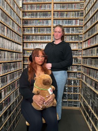 Odesa and Kate pose with serious faces in the music library, surrounded by CDs. Odesa is holding a capybara stuffy on her lap, like a child.