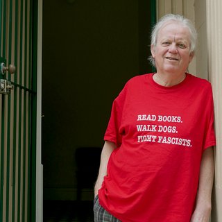 Stuart Coupe, host of Wild Card on Tuesdays, stands at a doorway wearing a red graphic tee that reads 'read books, walk dogs, fight fascists'