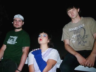 members of Ponzi Scheme shot against a fence with flash at night time. Kyle Innes in a earth green graphic t-shirt on the left, Vienna LaPalma McVernonin the centre wearing ultramarine fingerless gloves, and Thomas Harrison wearing a beige graphic t-shirt on the right.