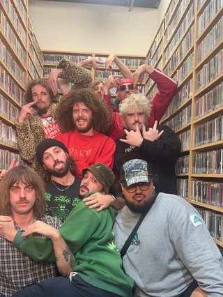 Nine members of Ferrari Party standing and sitting closely pulling gang signs with their hands surrounded by a library of CDs.