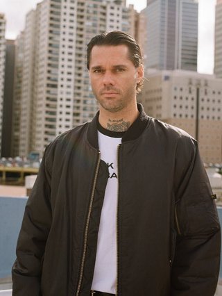 Shal in a black jacket and white shirt, looking forward with a serious expression