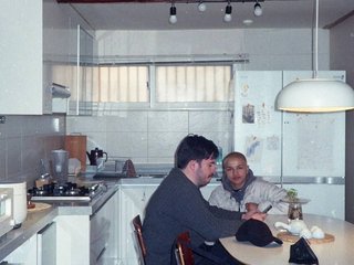 Pat sits at a dining table with his profile facing the camera. Rhea sits to his left facing the camera. They are in a brightly lit kitchen with white interiors and a wooden ceiling beam.