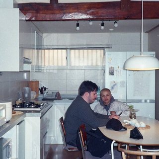 Pat sits at a dining table with his profile facing the camera. Rhea sits to his left facing the camera. They are in a brightly lit kitchen with white interiors and a wooden ceiling beam.