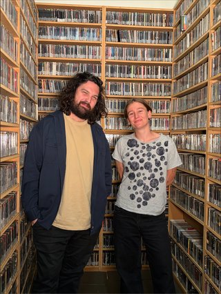 Blain, a man with long, curly dark hair, a yellow t shirt and navy blue jacket, and Gabs, a shorter white femme person with short blonde hair wearing a grey shirt with black circles and black pants, stand in front of floor-to-ceiling shelves of CDs. Blain has his hands in his pockets and Gabs has her hands behind her back. They both look at the camera, smiling slightly.