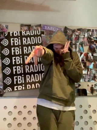 Jim wearing an olive green tracksuit in the fbi radio studio hitting a gyaru peace pose.