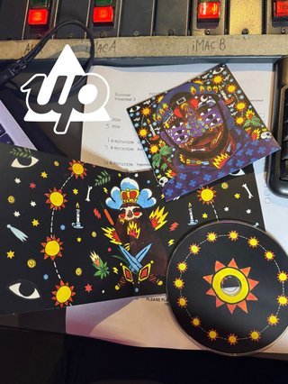 a photo of the cd 99.9%, Kaytranadas debut album. it is open and showcases the artwork; a sloth with symbols around him. the cd is in the bottom left corner; black with suns all over it and the lyric book in the top right which is a photo of the album cover