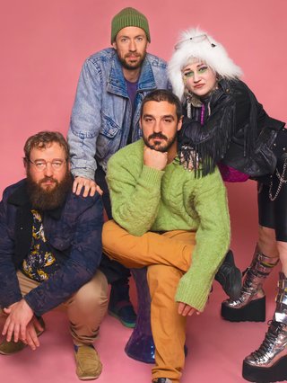 All four members of Hiatus Kaiyote leaning into each other both crouching and standing before a salmon pink backdrop.