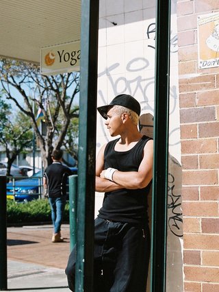 Fabz is seen leaning against a wall, looking away and biting his lips. He is captured in the reflection of a mirror. Fabz wears a black cap to the side, a black singlet and dark denim jorts.