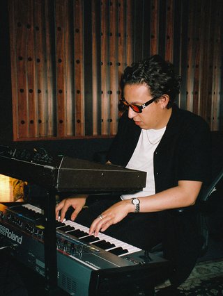 Nicolas Mendoza wears sits as he plays keys in a darkened room with wood panels behind him. He wears a white t-shirt, black short sleeve button up shirt, and sunglasses.