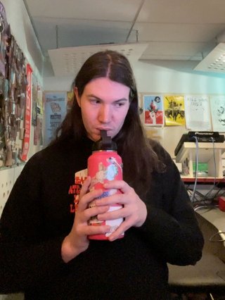 Jim sitting in the fbi studio, sipping from their water bottle which is pink and covered in stickers.
