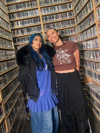 Two people standing in front of shelves of cds. Ecca has blue hair, wearing a black jacket and a blue dress. Bri is wearing a small brown tshirt and a black skirt.