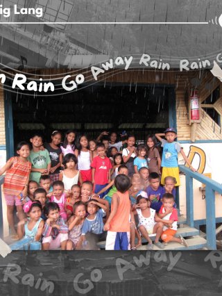 A group photo of kids singing “Rain Rain Go away,” standing on the steps of a hut superimposed on top of some damaged rubble